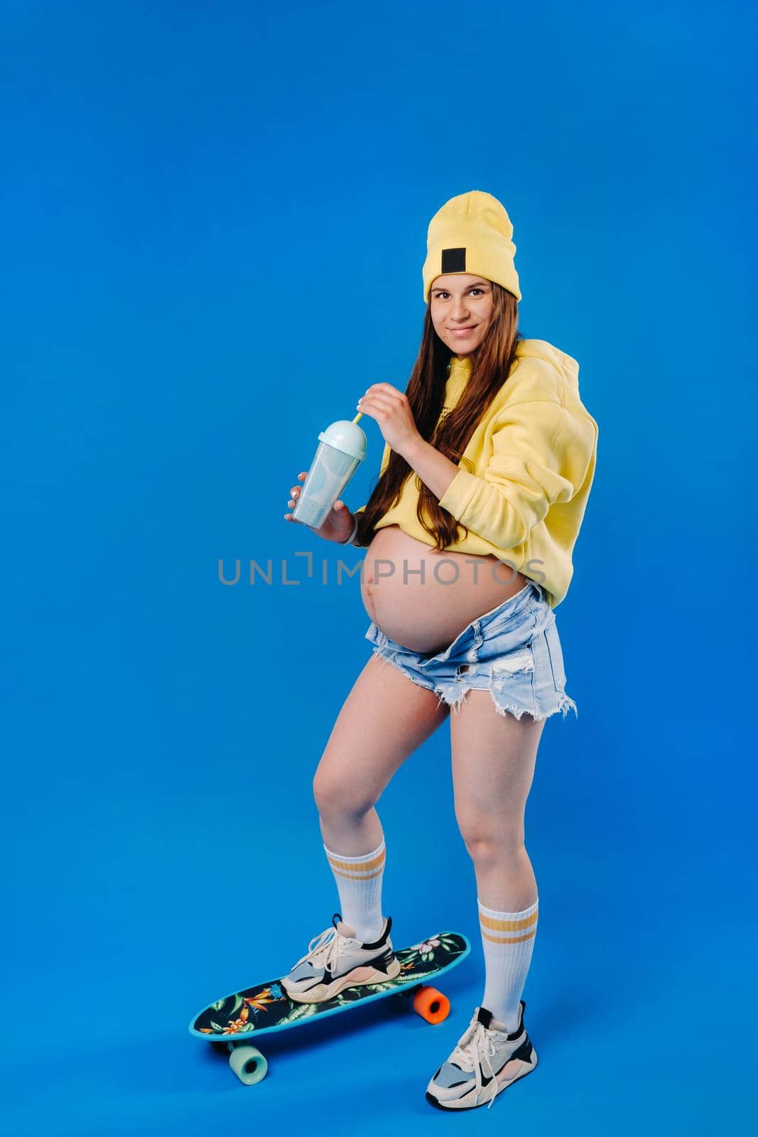a pregnant girl in yellow clothes with a glass of juice rides a skateboard on a blue background.