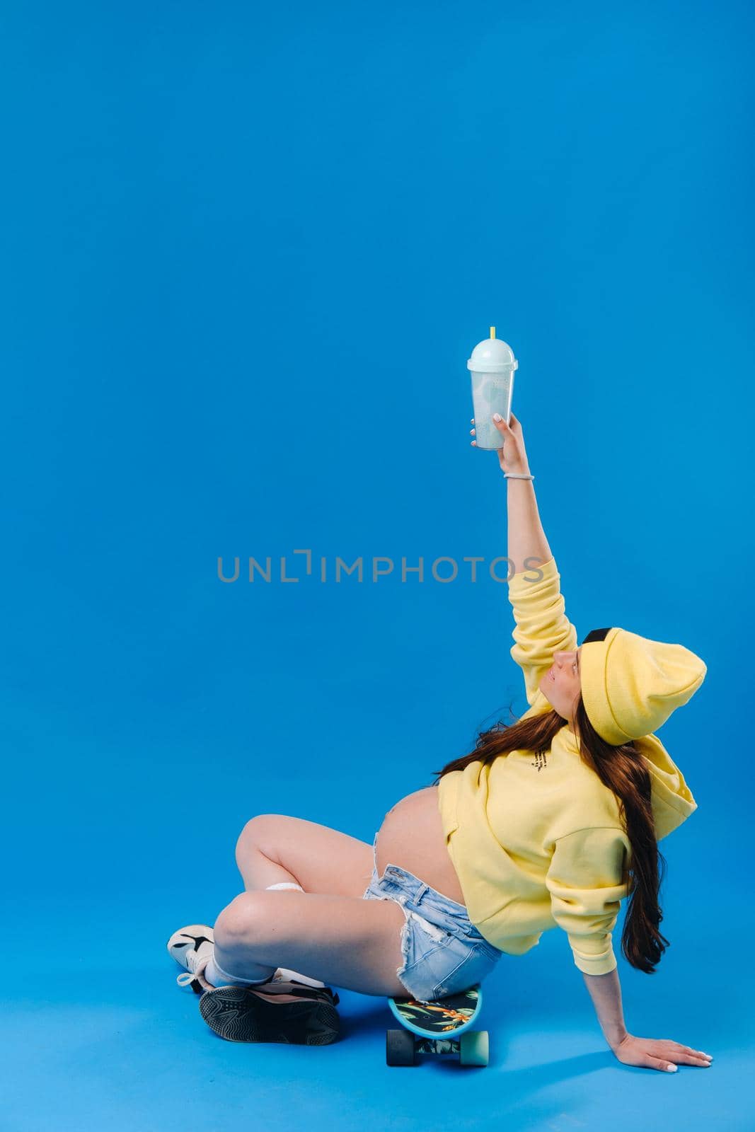 a pregnant girl in yellow clothes with a glass of juice sits on a skateboard on a blue background.