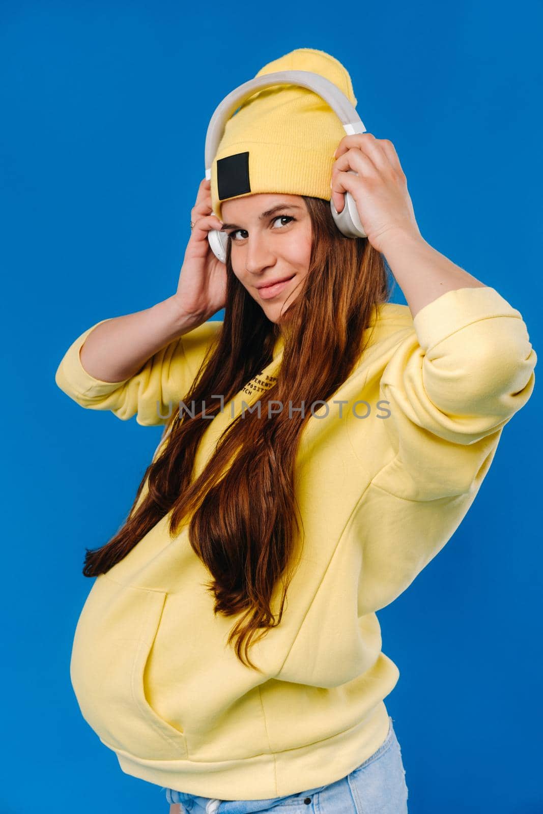 A pregnant girl in a yellow jacket and headphones stands on a blue background by Lobachad