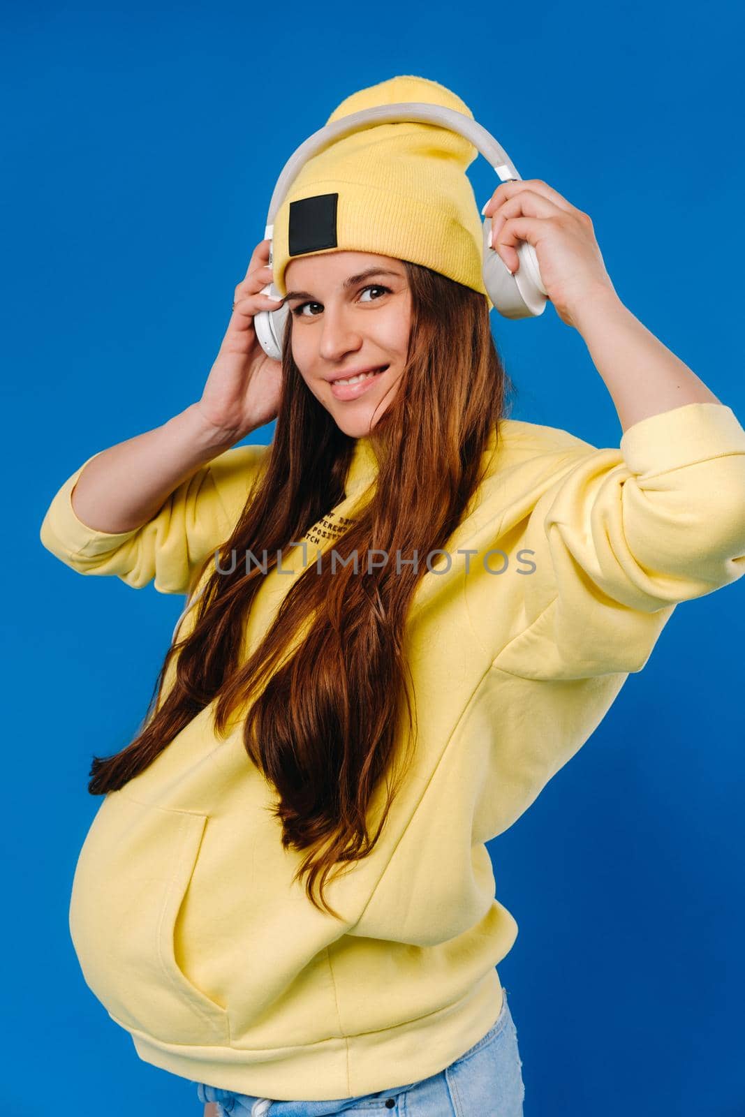 A pregnant girl in a yellow jacket and headphones stands on a blue background by Lobachad