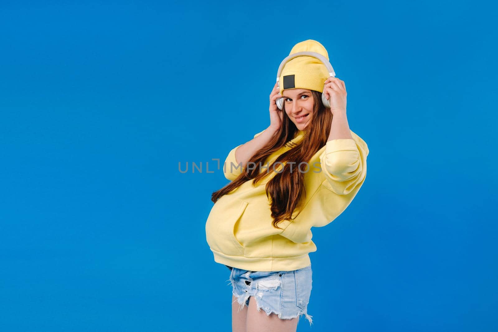 A pregnant girl in a yellow jacket and headphones stands on a blue background.
