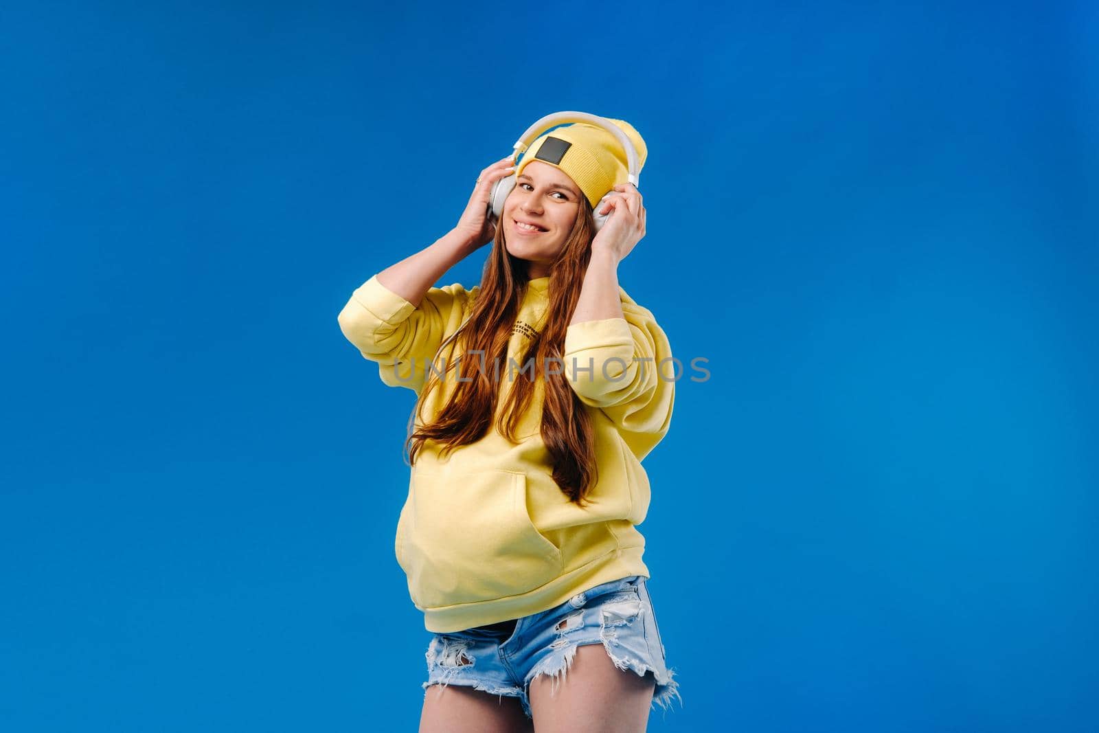 A pregnant girl in a yellow jacket and headphones stands on a blue background.