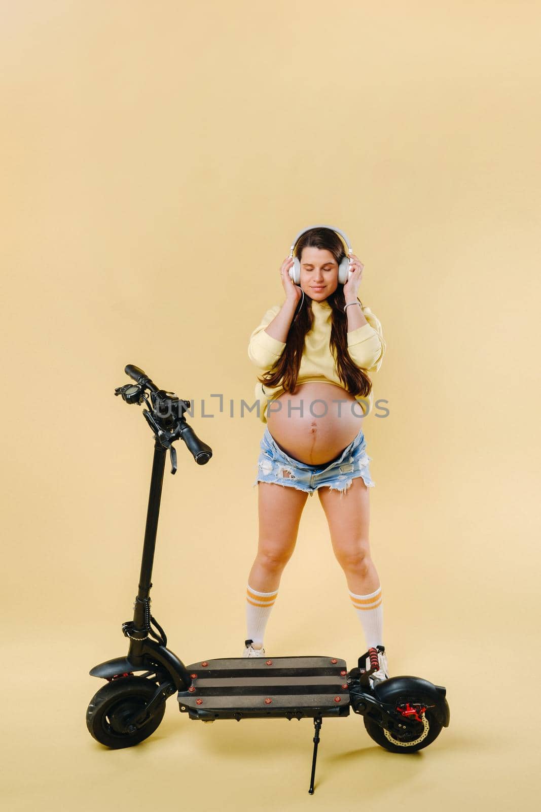 Pregnant girl in yellow clothes and headphones on an electric scooter on an isolated yellow background by Lobachad