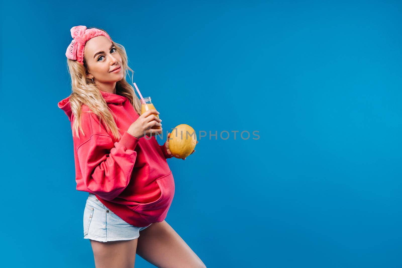 pregnant girl in pink clothes with a bottle of juice and a melon on a blue background.