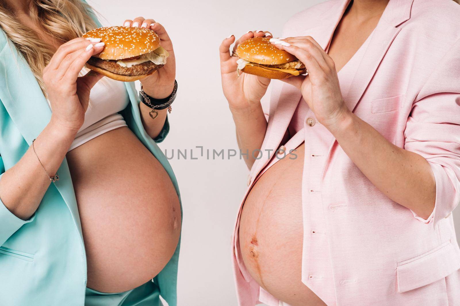 Two pregnant women in suits with hamburgers in their hands on a gray background.