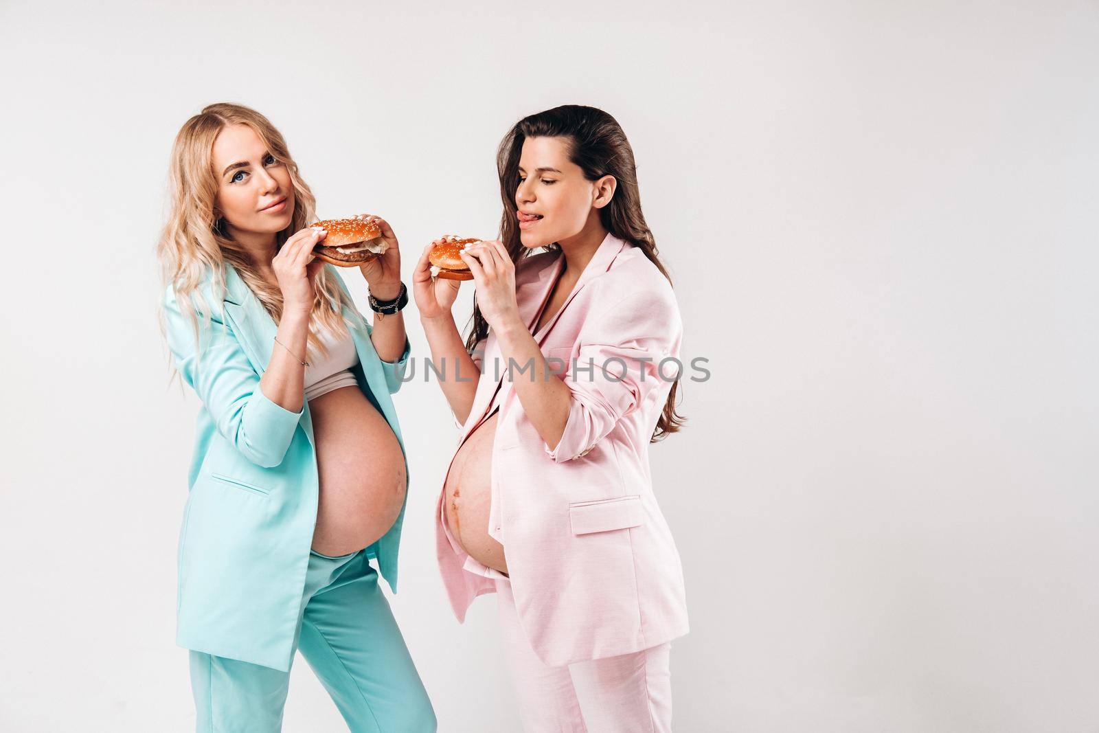 Two pregnant women in suits with hamburgers in their hands on a gray background.
