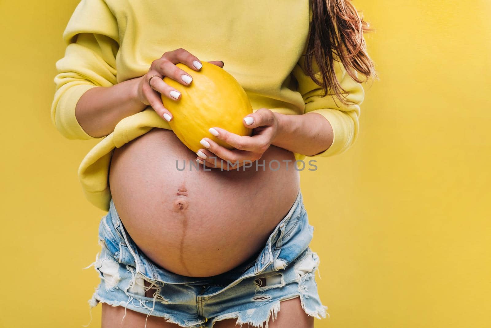 a pregnant girl with a yellow jacket stands with a melon in her hands on a yellow isolated background.