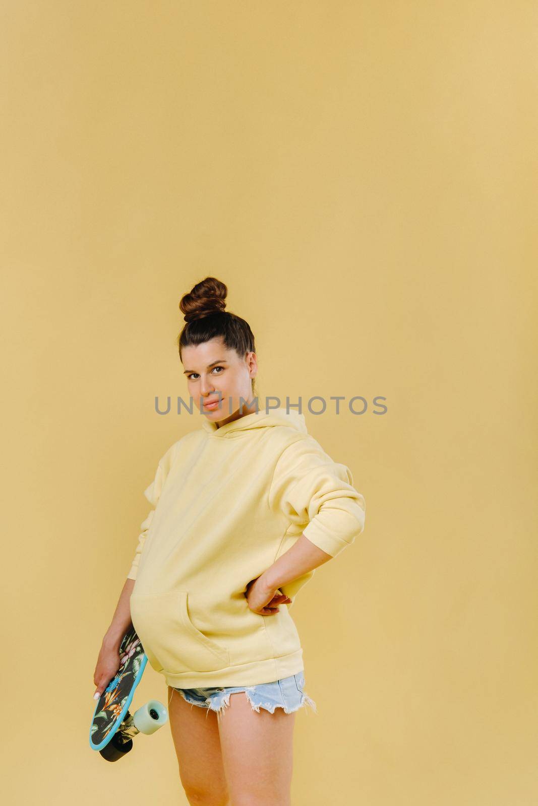 Pregnant girl in a yellow jacket with a skateboard in her hands on a yellow background.