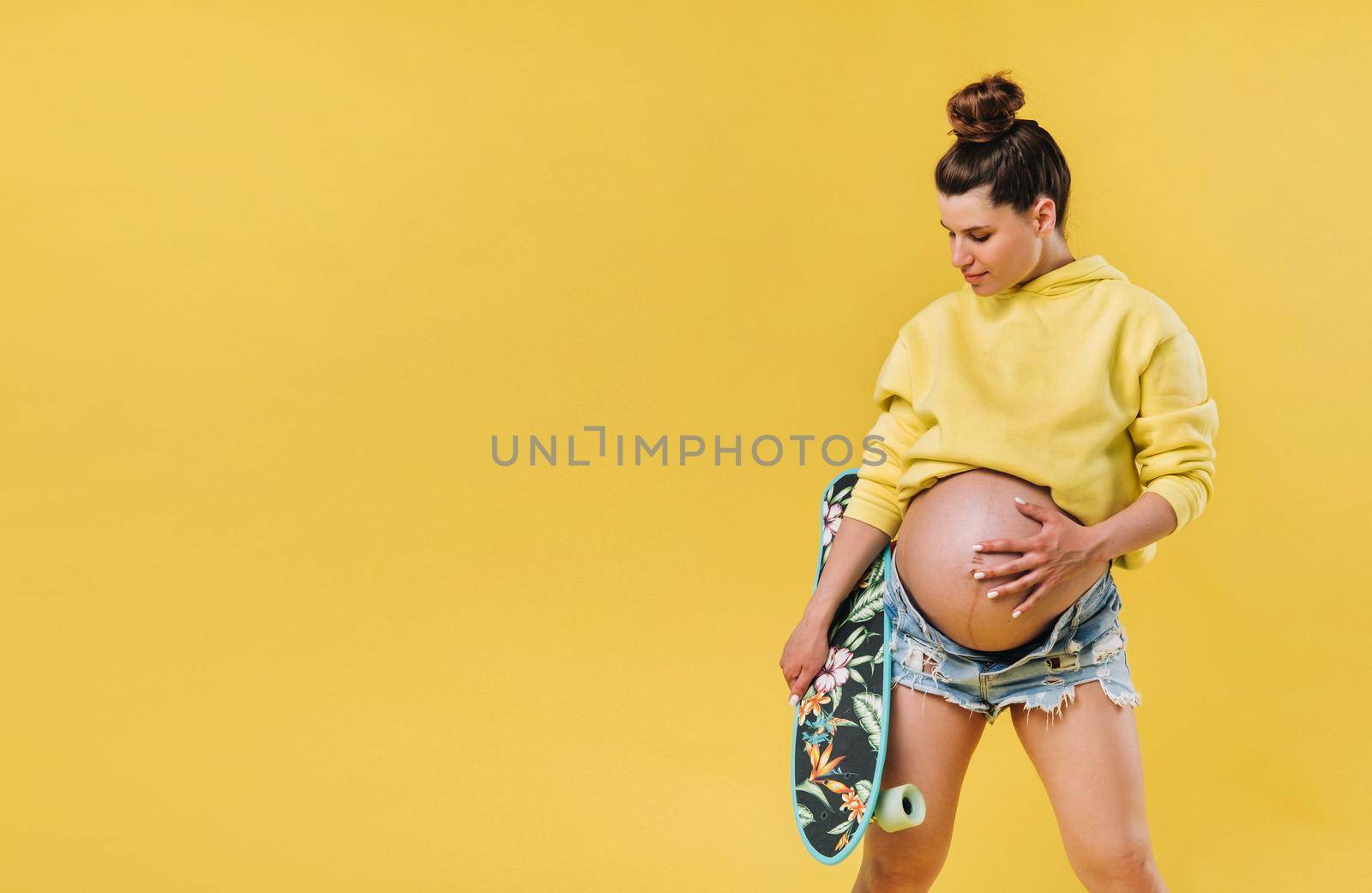Pregnant girl in a yellow jacket with a skateboard in her hands on a yellow background.