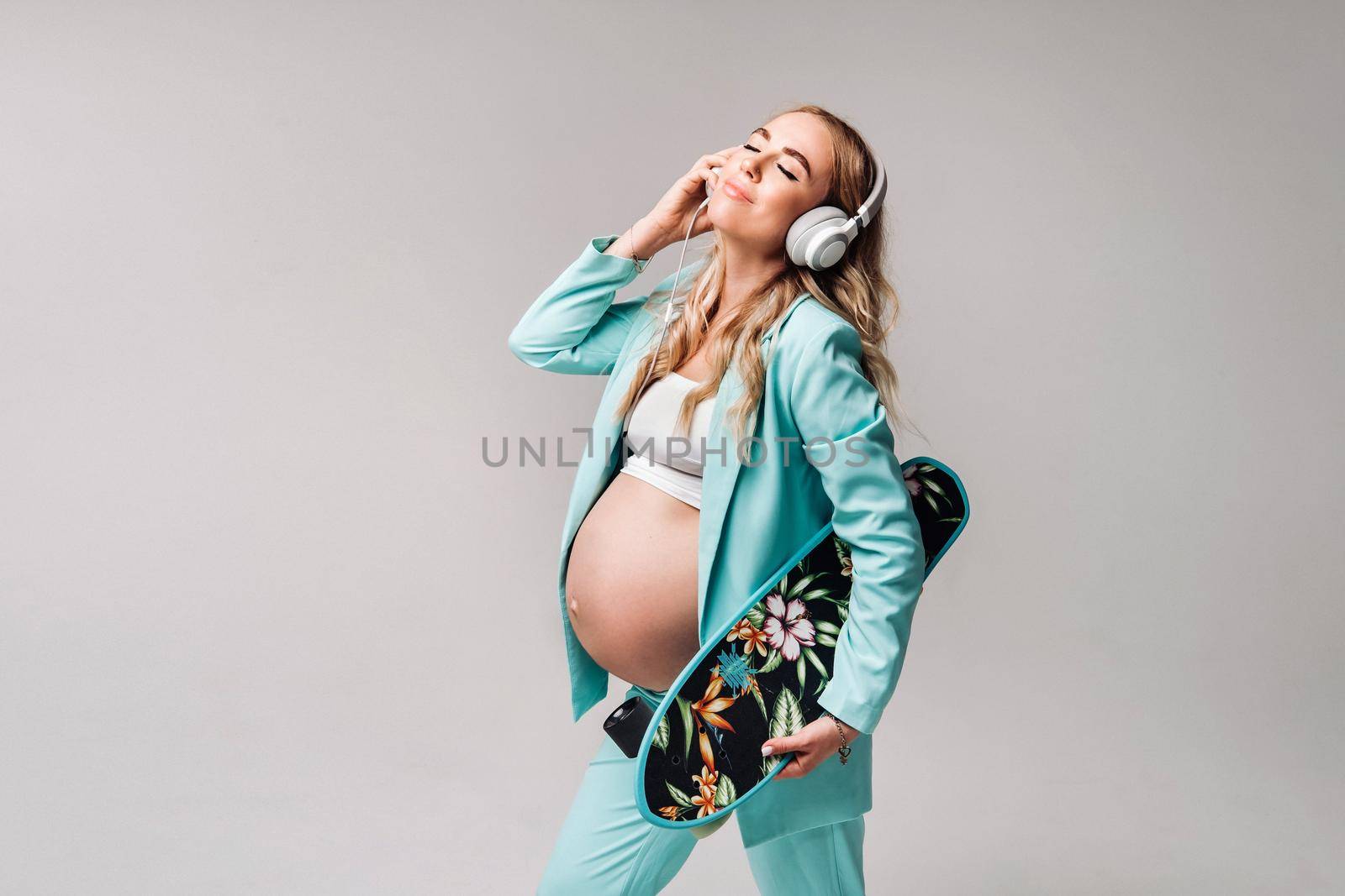 A pregnant girl in a turquoise suit with a skateboard in her hands and headphones stands on a gray background.