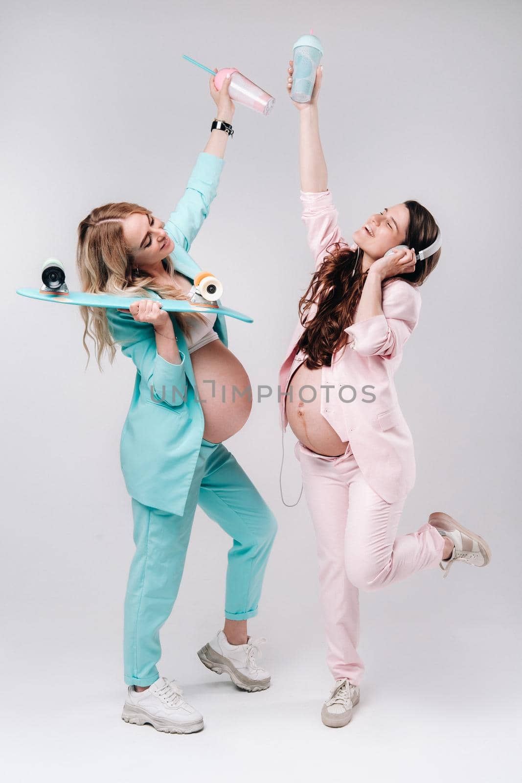 Two pregnant girls in turquoise and pink suits with glasses of juice, a skate and headphones stand on a gray background by Lobachad