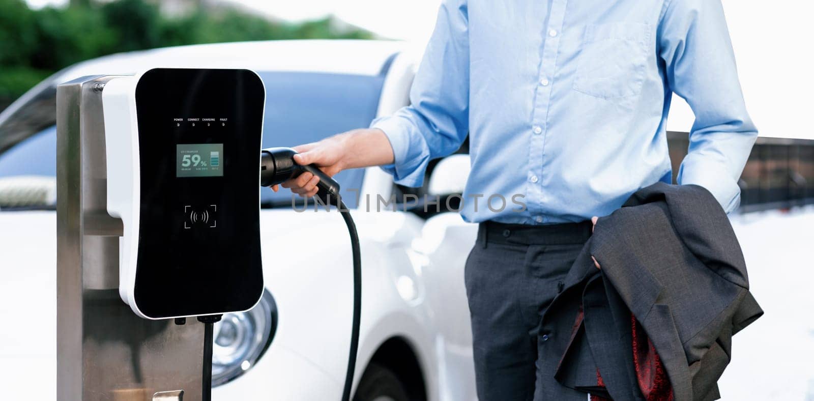 Closeup progressive suit-clad businessman with his electric vehicle recharge his car on public charging station in modern city with power cable plug and renewable energy-powered electric vehicle.