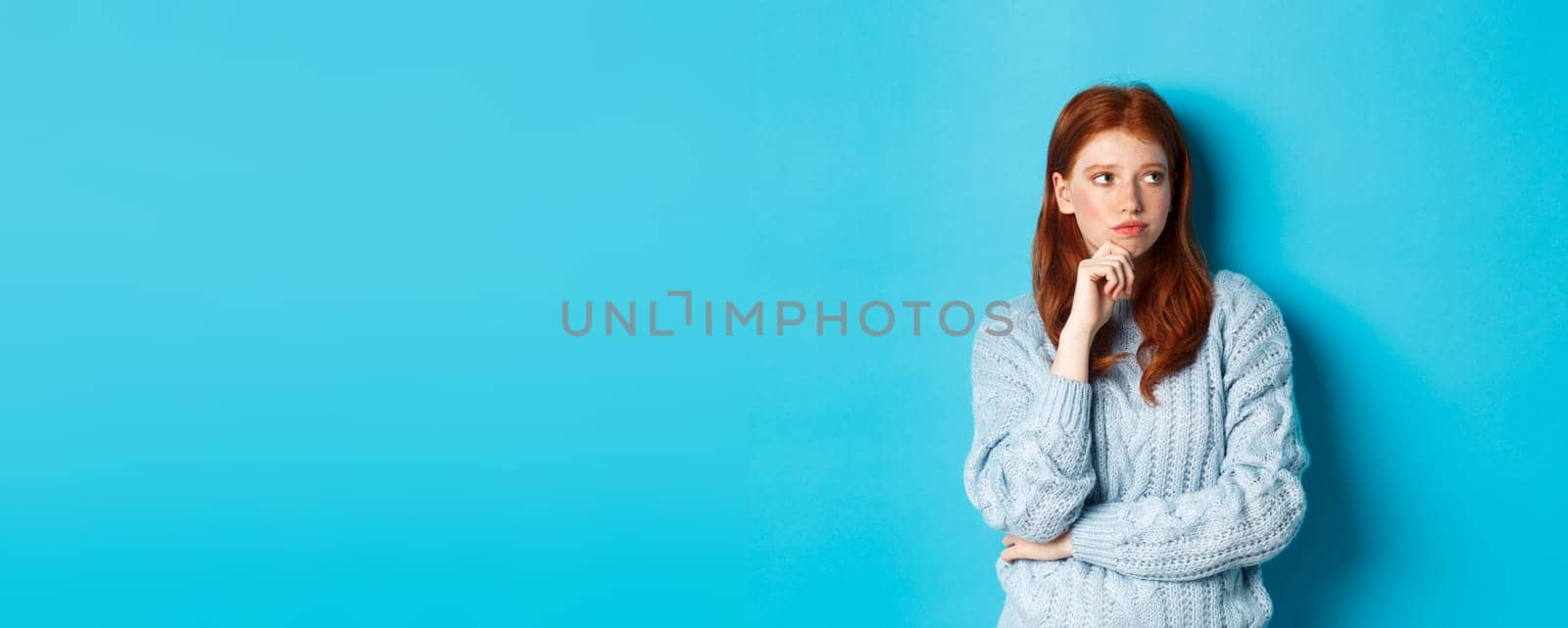 Thoughtful and upset redhead girl looking right, pondering solution, standing in sweater against blue background by Benzoix