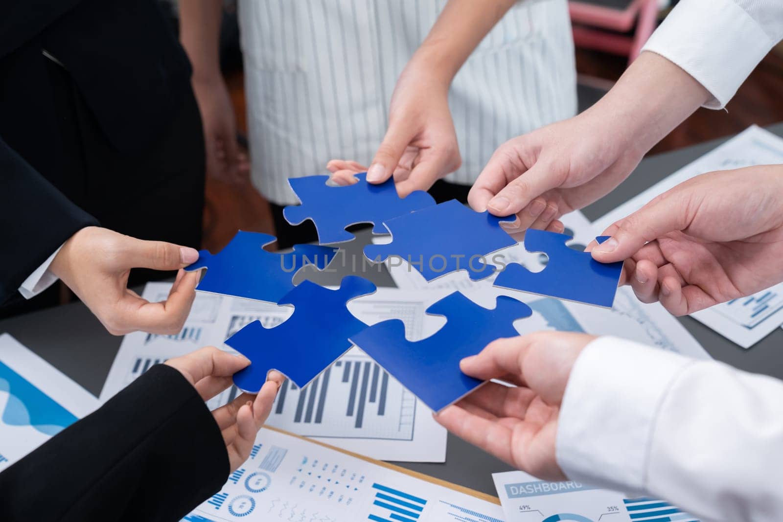 Closeup top view business team of office worker putting jigsaw puzzle together over table filled with financial report paper in workplace with manager to promote harmony concept in meeting room.