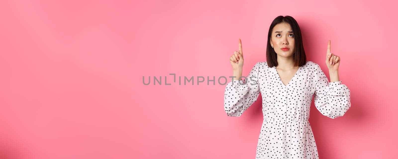 Worried and concerned asian girl in cute dress frowning, pointing and looking up with sad face, standing over pink background by Benzoix