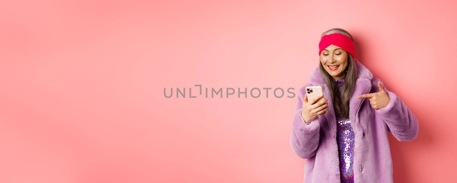 Online shopping and fashion concept. Cool asian senior woman checking out promotion on mobile phone, pointing finger at smartphone screen and smiling pleased, pink background by Benzoix