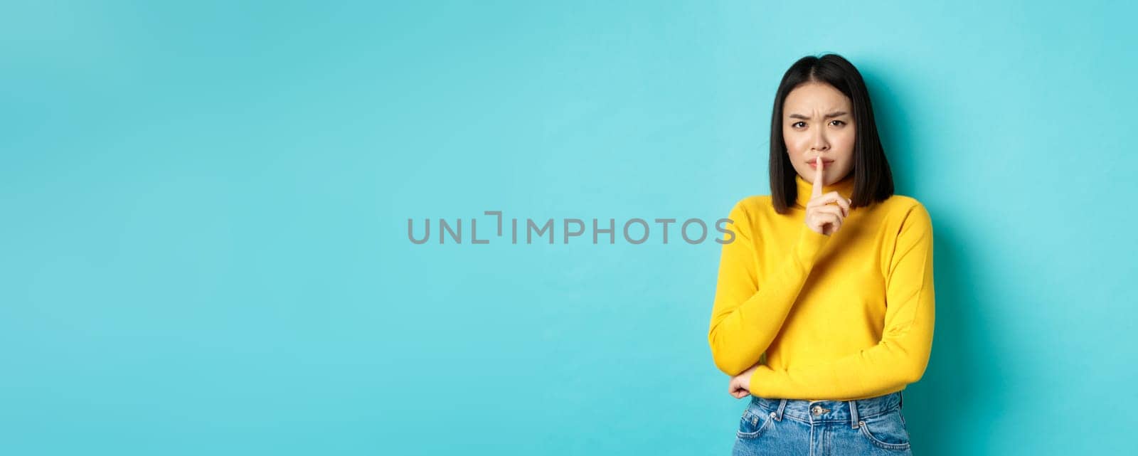 Disappointed asian woman telling to be quiet, scolding loud person with hush gesture, shushing at camera and frowning upset, standing over blue background.