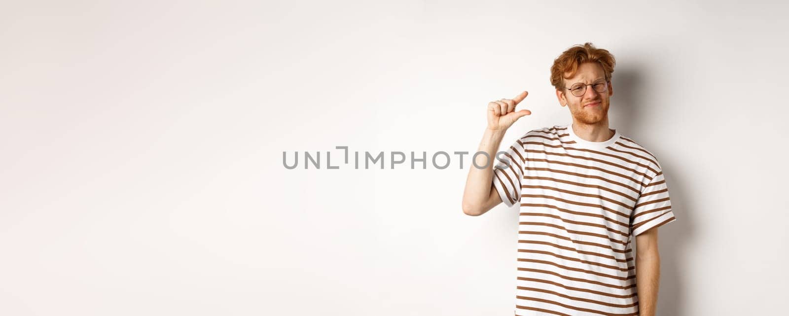 Disappointed young man with red hair and beard, wearing glasses and showing small size, little thing, standing over white background.