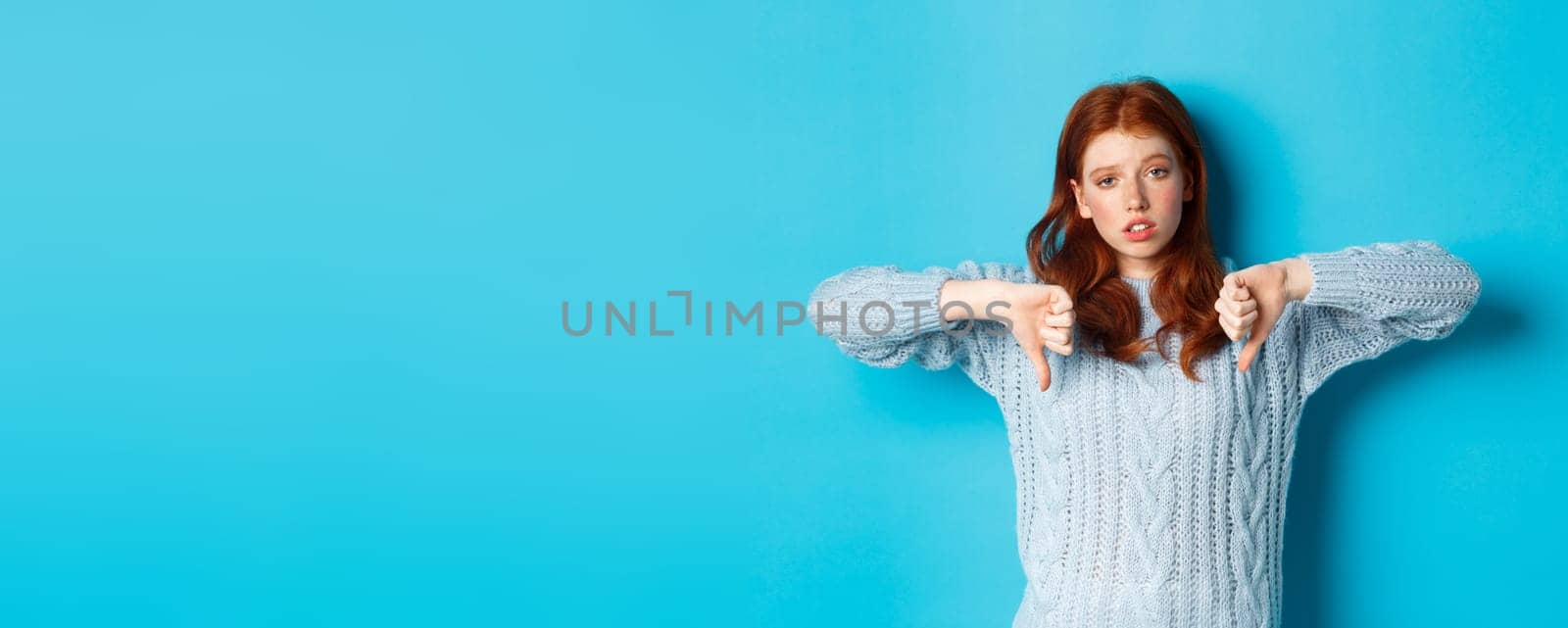 Bored and skeptical redhead girl showing thumbs down, looking unamused and uninterested, standing over blue background.