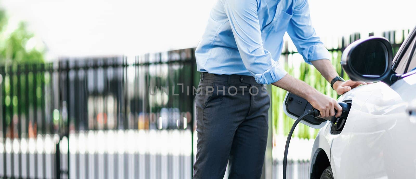 Closeup progressive man holding EV charger plug from public charging station for electric vehicle with background of residential building as concept eco-friendly sustainability energy car concept.