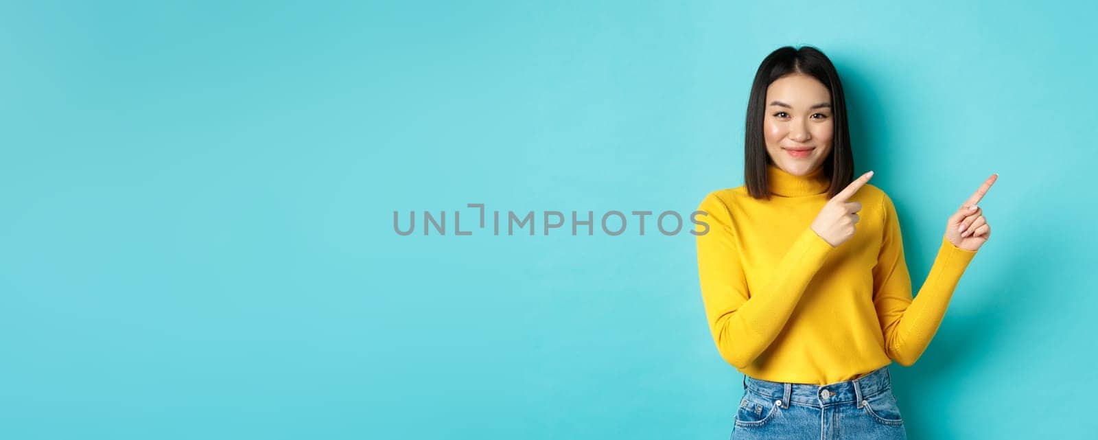 Shopping concept. Attractive young asian woman with perfect skin, wearing trendy yellow sweater and jeans, pointing fingers right and smiling, showing advertisement.