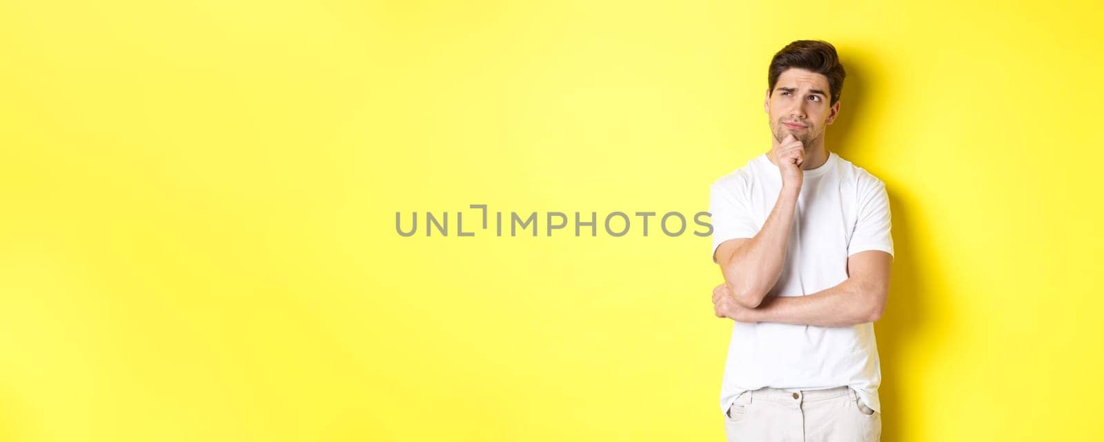 Portrait of young male model thinking, looking at upper left corner and making choice, standing near copy space, yellow background.