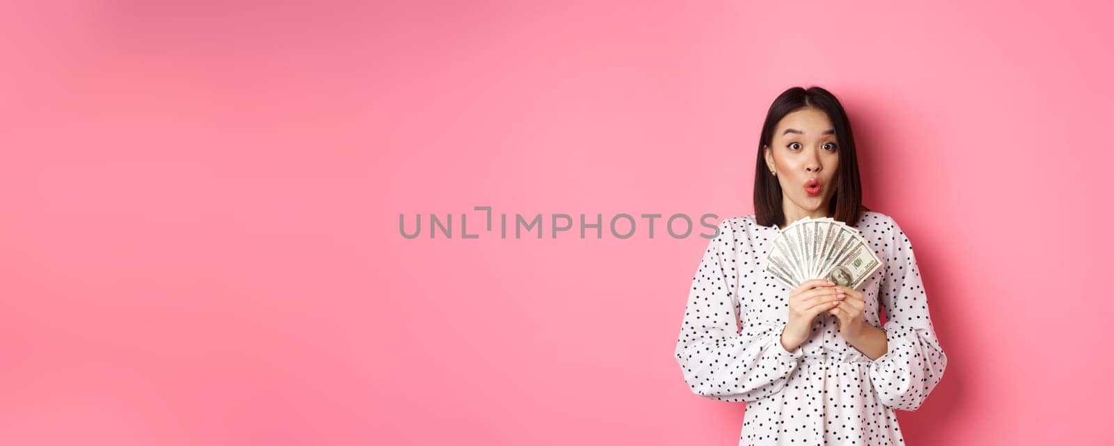 Shopping concept. Excited asian woman holding money, gasping amazed and staring at camera, standing over pink background by Benzoix