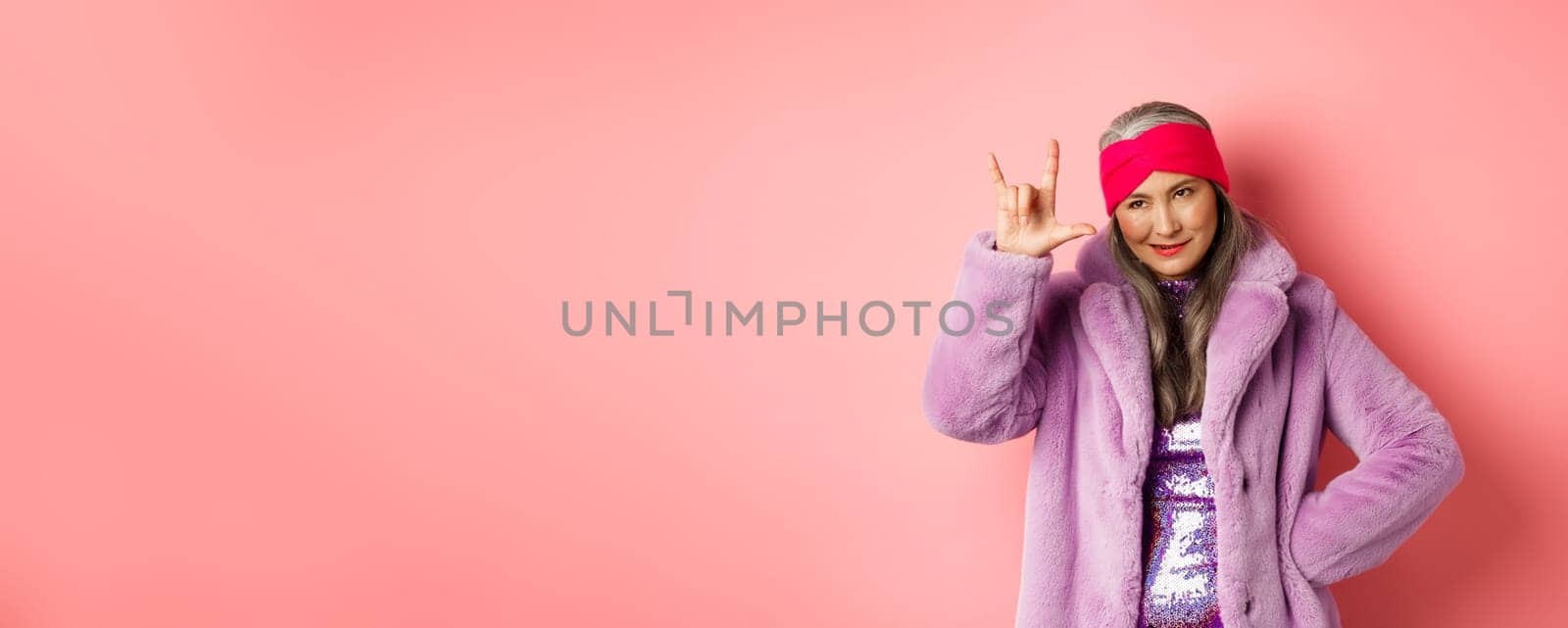 Funny and cool asian senior woman showing rock n roll gesture, looking sassy, standing over pink background by Benzoix