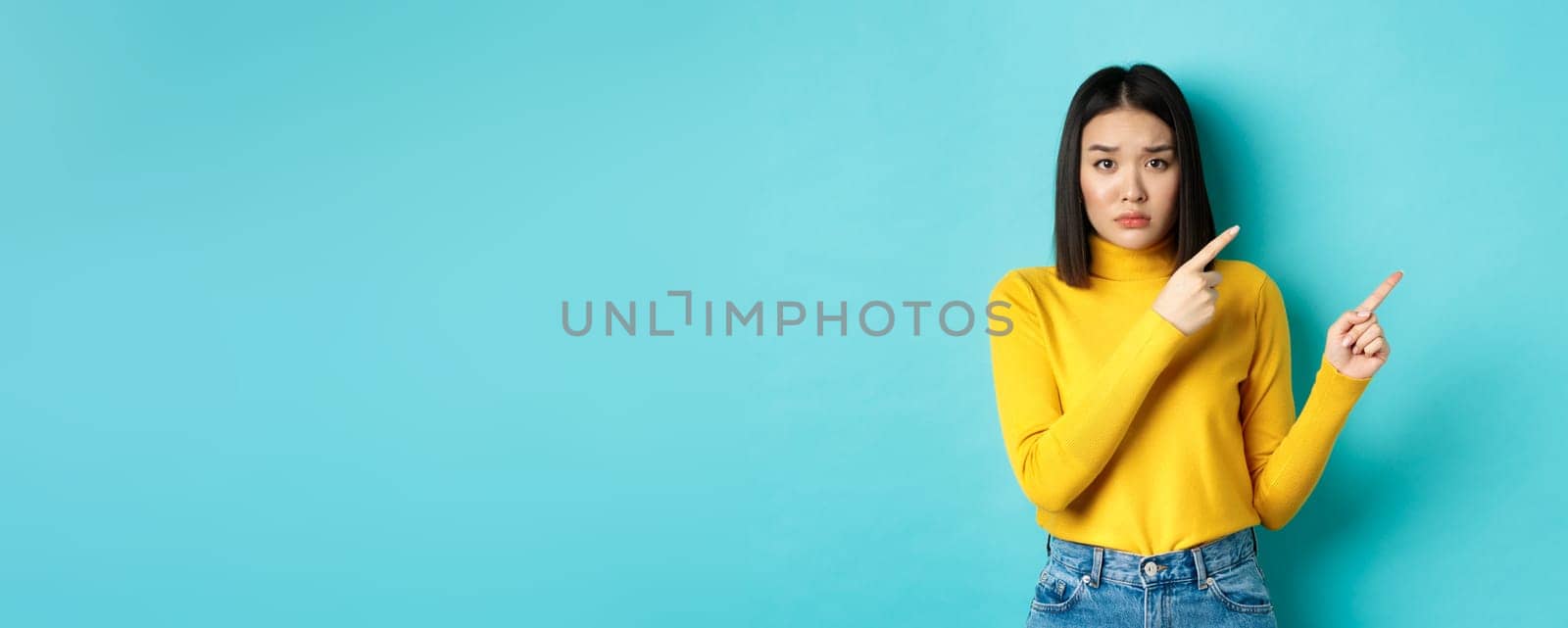 Shopping concept. Disappointed korean girl looking gloomy, asking to buy this, pointing fingers at upper right corner and staring sad at camera, blue background by Benzoix