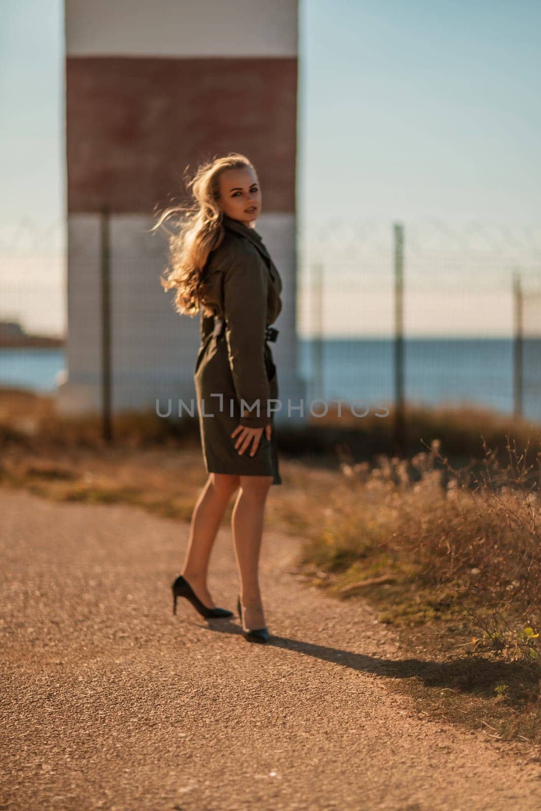 Portrait blonde sea cape. A calm young blonde in a khaki raincoat stands on the seashore against the backdrop of a lighthouse