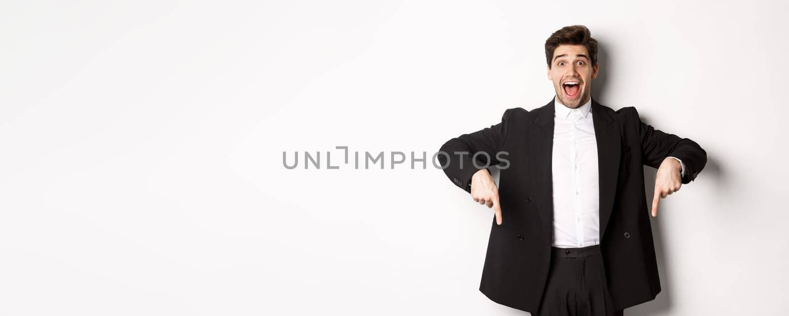 Portrait of super happy man showing christmas promo offer, pointing fingers down and smiling amazed, wearing formal party suit, standing over white background by Benzoix