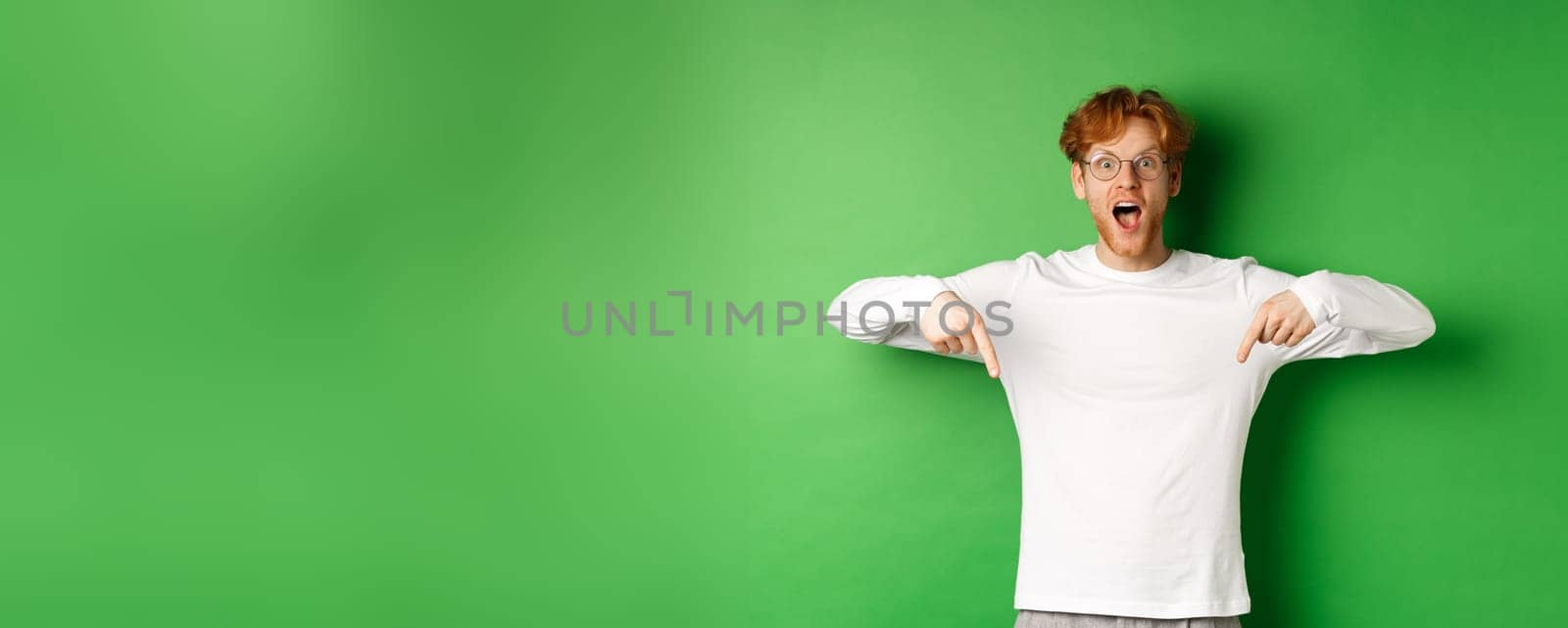 Surprised and excited young redhead man scream of joy and amazement, checking out awesome promo, pointing fingers down, standing over green background.