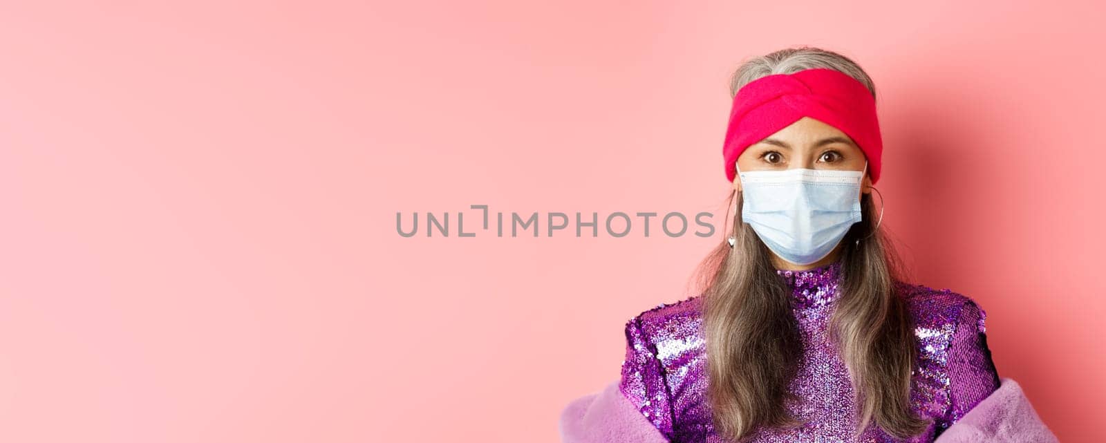 Covid-19, virus and social distancing concept. Close-up of stylish asian senior woman in medical mask and shiny dress looking at camera, standing over pink background.