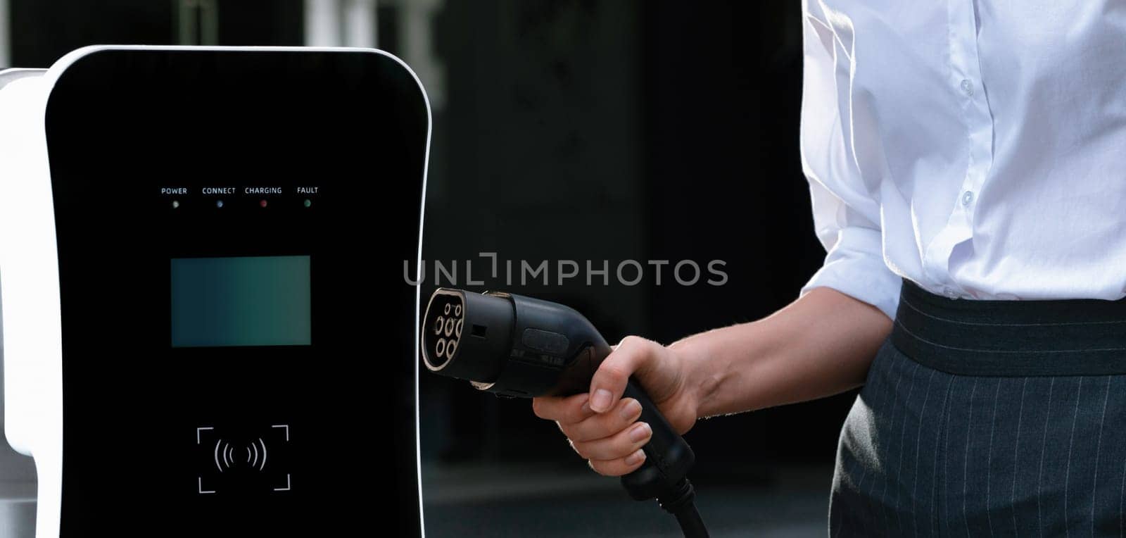 Focus EV charger plug and electric car at public charging station with blur progressive businesswoman holding charger and apartment condo building in background. Eco friendly electric vehicle concept.