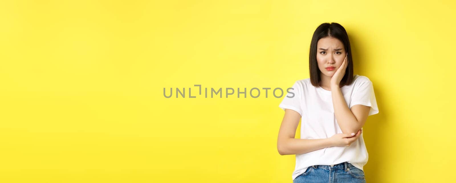 Sad asian woman touching cheek and frowning, having toothache, need see dentist, standing gloomy against yellow background by Benzoix