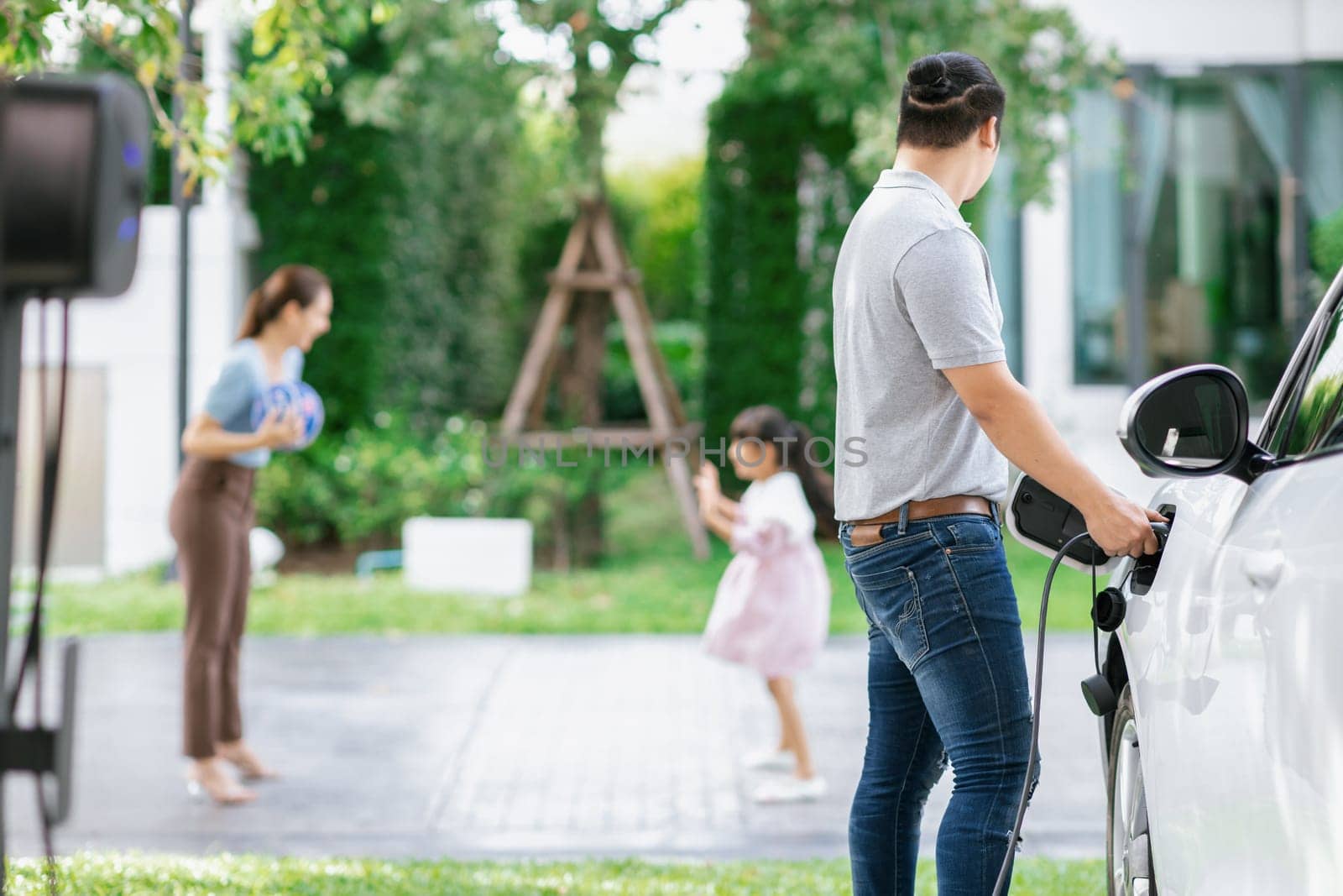 Focus image of progressive man charging electric car from home charging station with blur mother and daughter playing together in the background.