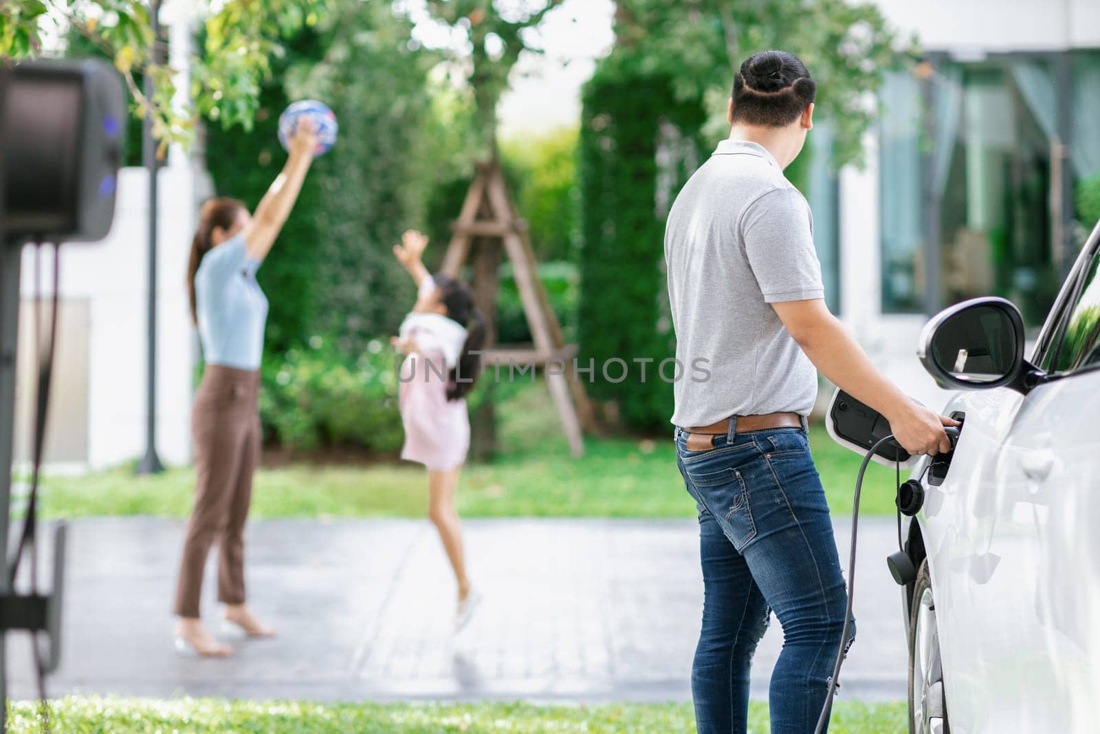 Focus image of progressive man charging electric car from home charging station with blur mother and daughter playing together in the background.