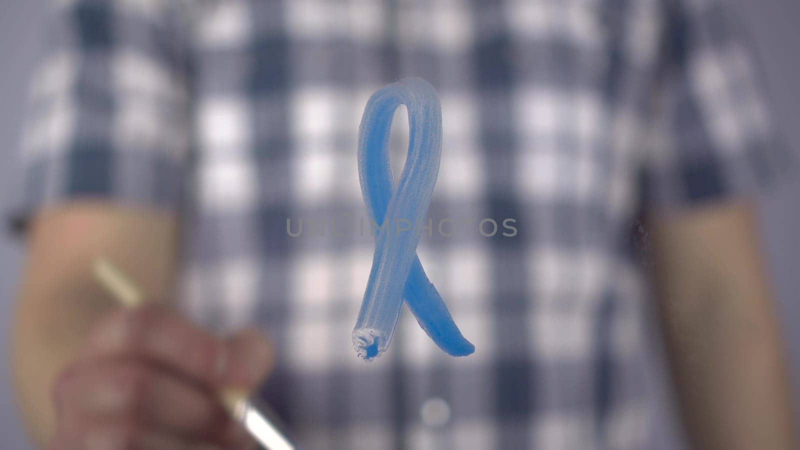 A man draws a blue ribbon on the glass in support of cancer patients. A young man paints with a brush the symbol of prostate cancer. Close-up. 4k