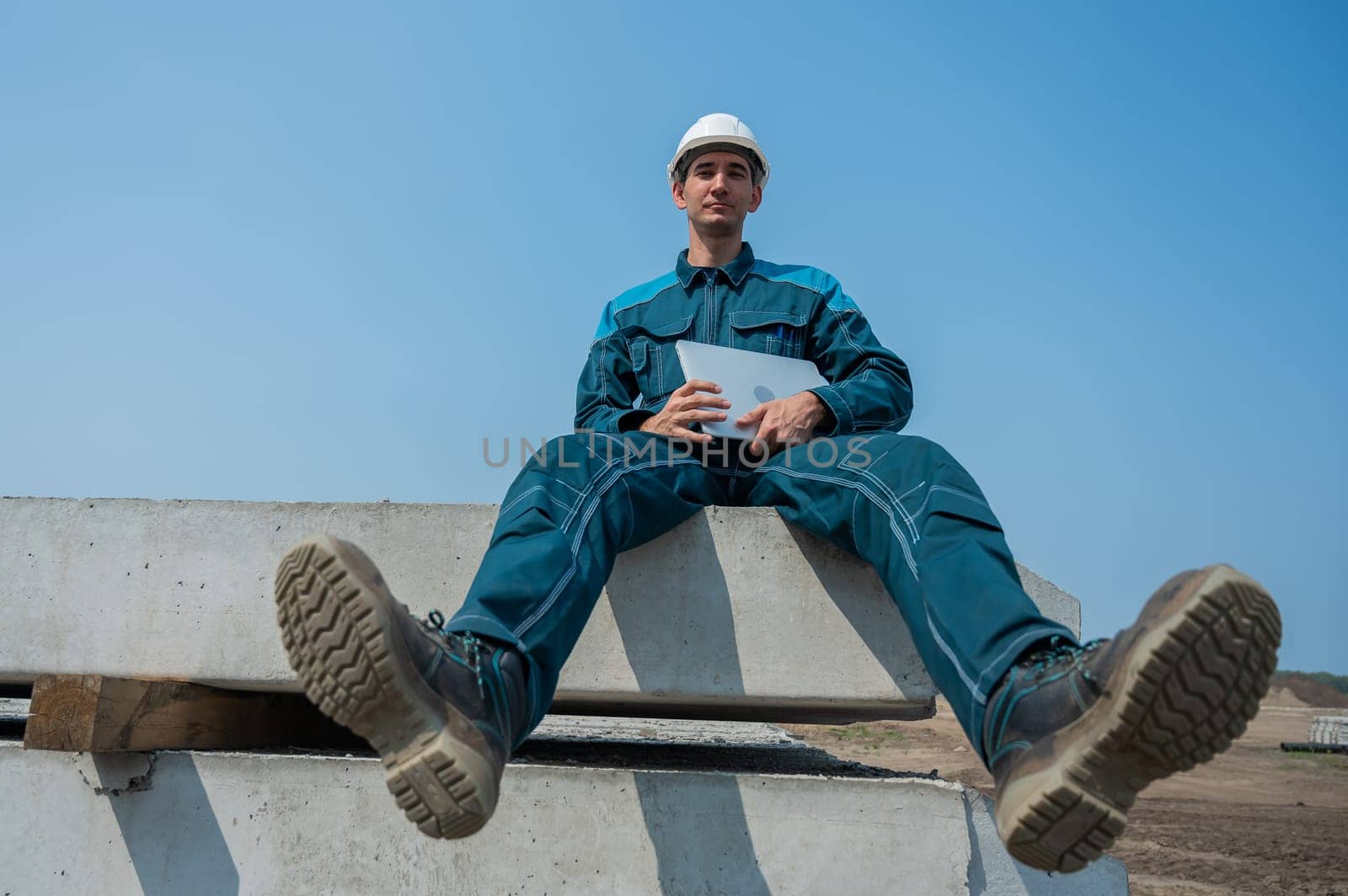 Caucasian male builder in hardhat sits on floor slabs and uses laptop at construction site. by mrwed54