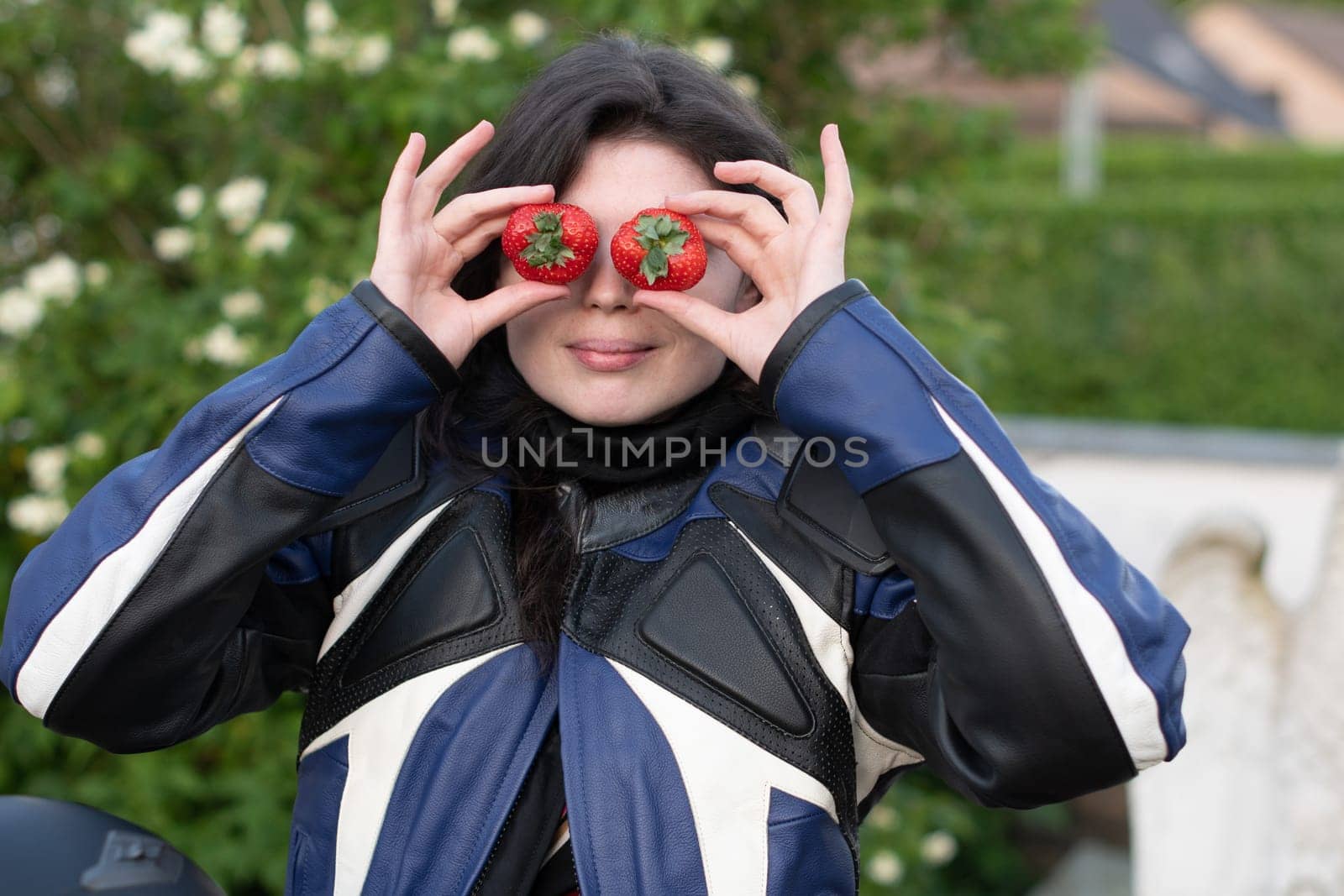 beautiful young woman motobiker in black leather pants, black boots and a black helmet, in a blue jacket on a red honda motorcycle, put strawberries to her eyes. High quality photo