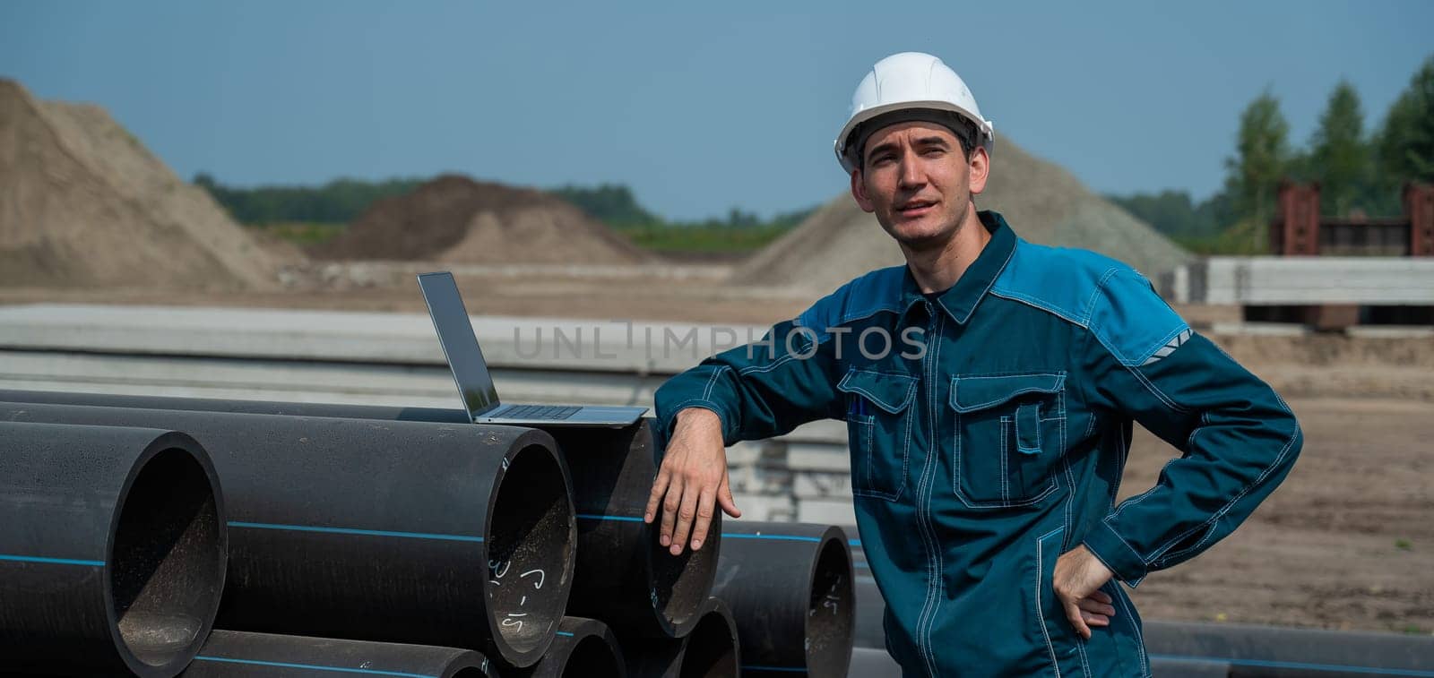 Caucasian male builder in a hard hat stands near the pipes and uses a laptop at a construction site. by mrwed54