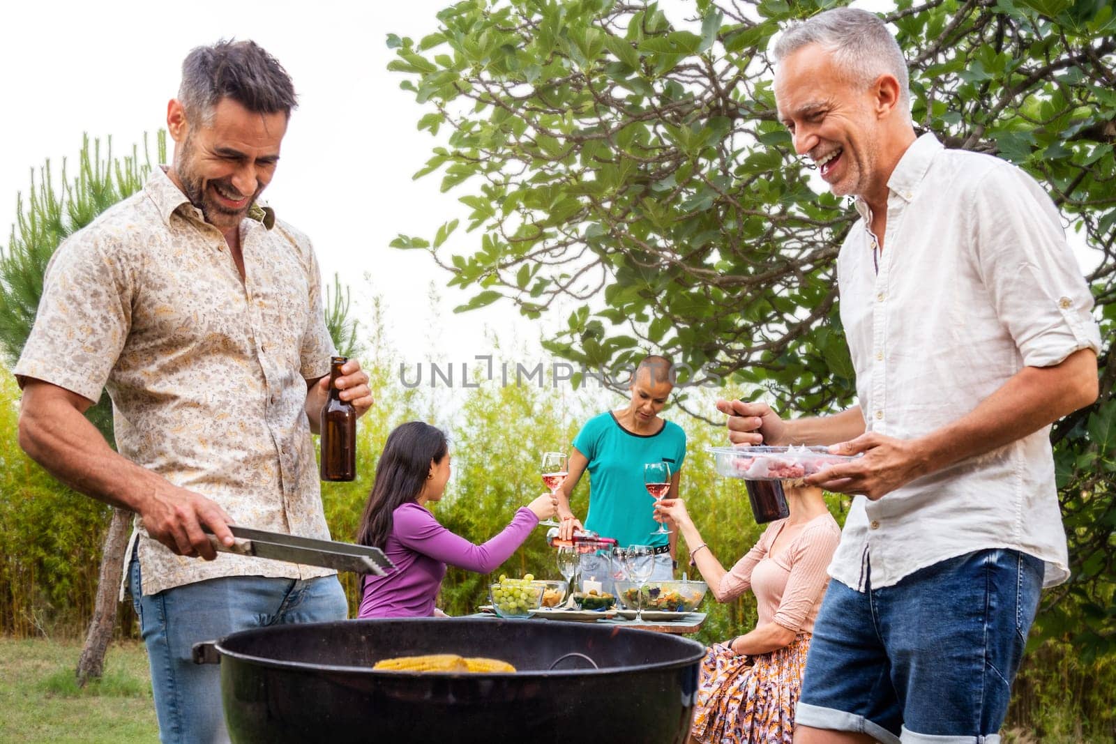 Two men cooking food on grill. Outdoor garden barbecue party. Three women enjoying wine on background. Weekend activities concept.