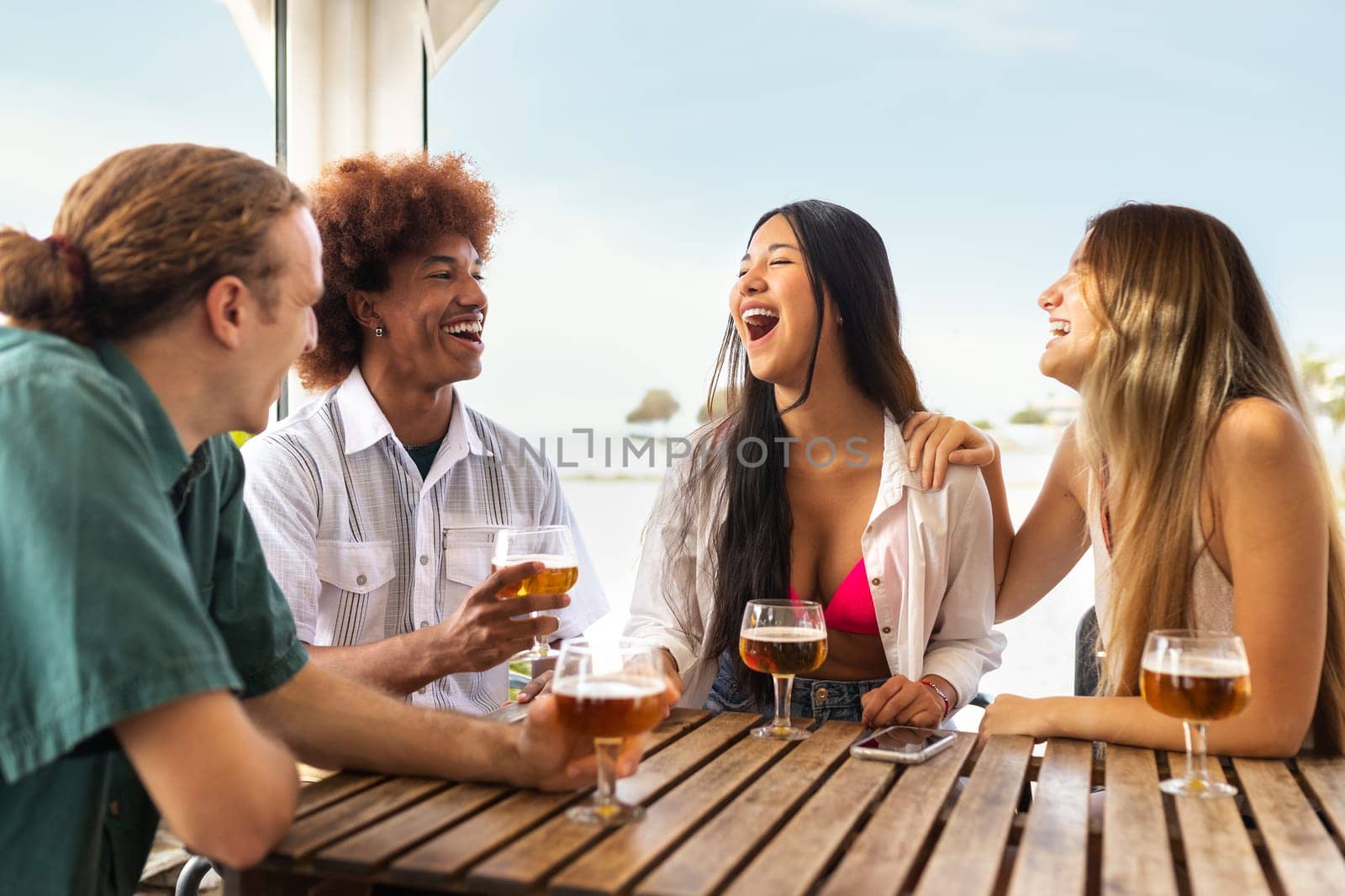 Group of multiracial young friends laughing, talking and having fun at beach bar drinking beers together. Lifestyle concept.