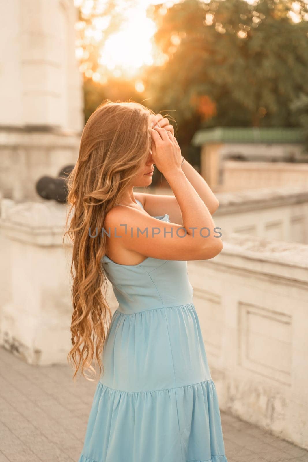 Woman sunset blue dress. Portrait of a woman with long hair and a blue dress against the backdrop of the setting sun and a white building. Lifestyle, walking around the city by Matiunina