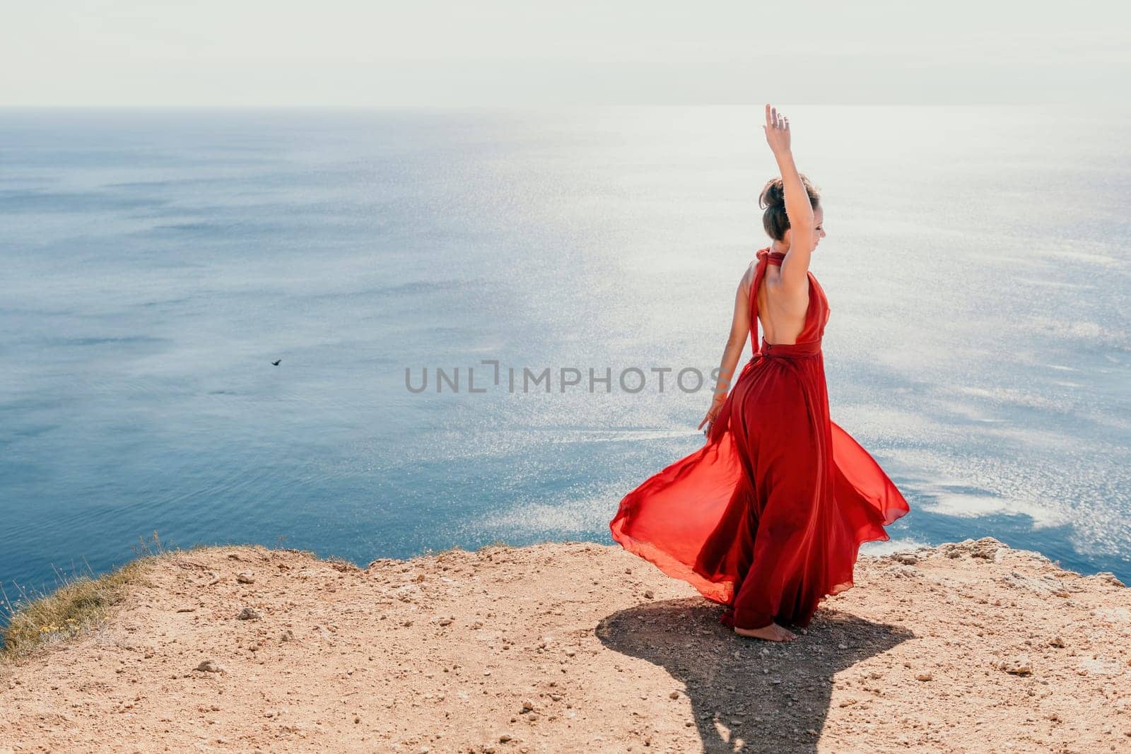 Side view a Young beautiful sensual woman in a red long dress posing on a rock high above the sea during sunrise. Girl on the nature on blue sky background. Fashion photo.