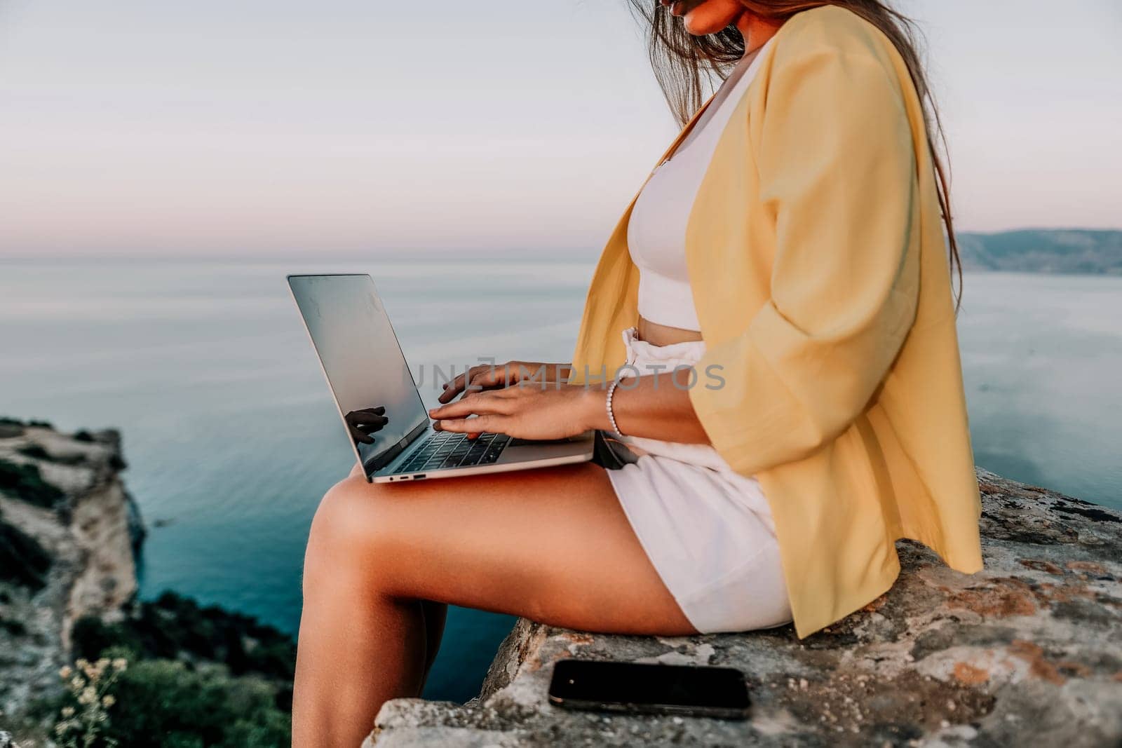 Digital nomad, Business woman working on laptop by the sea. Pretty lady typing on computer by the sea at sunset, makes a business transaction online from a distance. Freelance, remote work on vacation by panophotograph