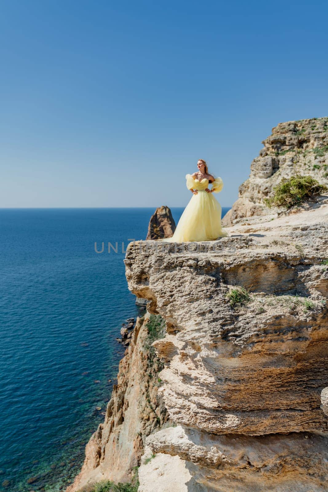 Woman in a yellow dress on the sea. Side view Young beautiful sensual woman in yellow long dress posing on a rock high above the sea at sunset. Girl in nature against the blue sky by Matiunina