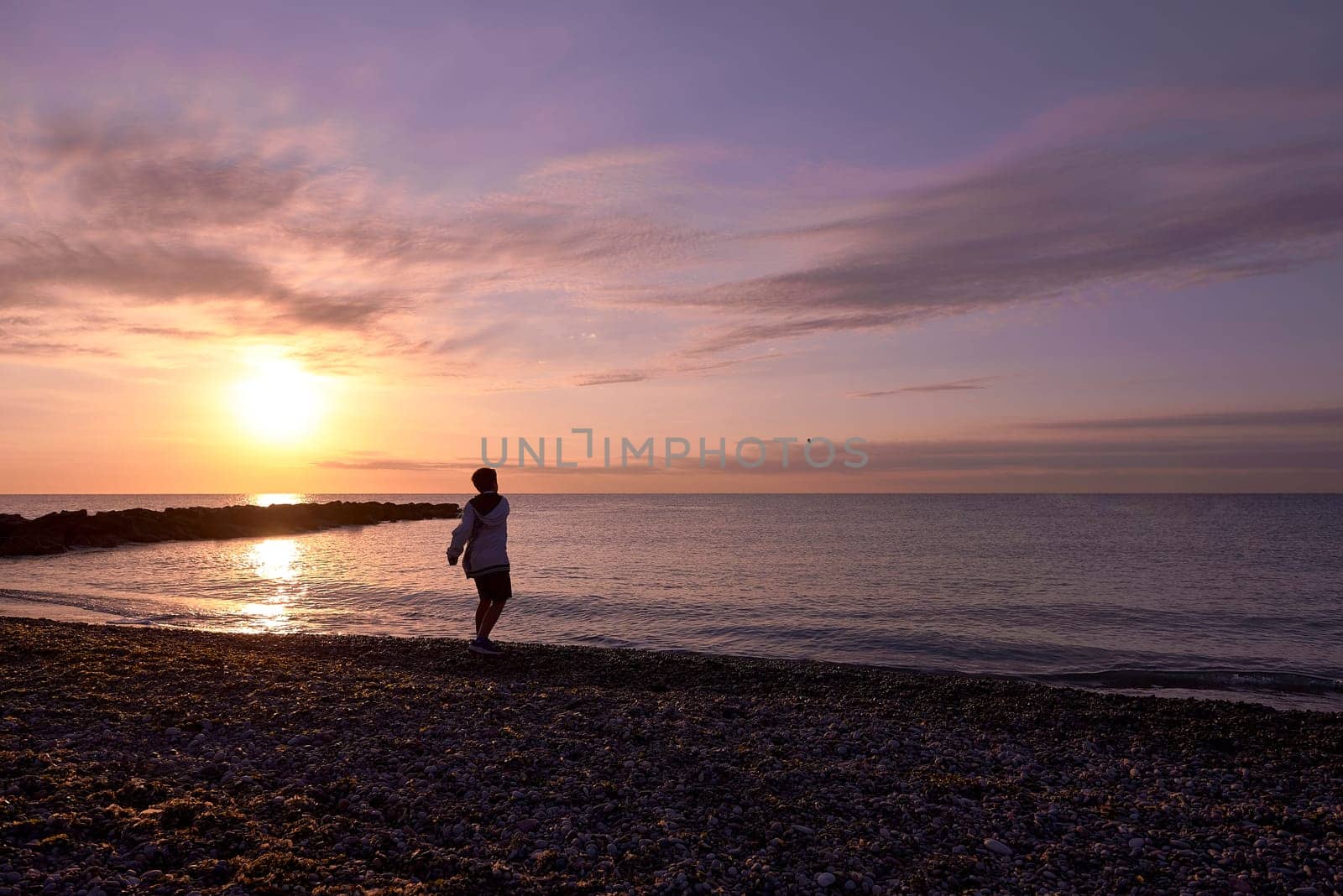 Boy playing pebble throwing on the beach at dawn. Unrecognisable, small pebble beach, sun, yellow and orange colours, life style