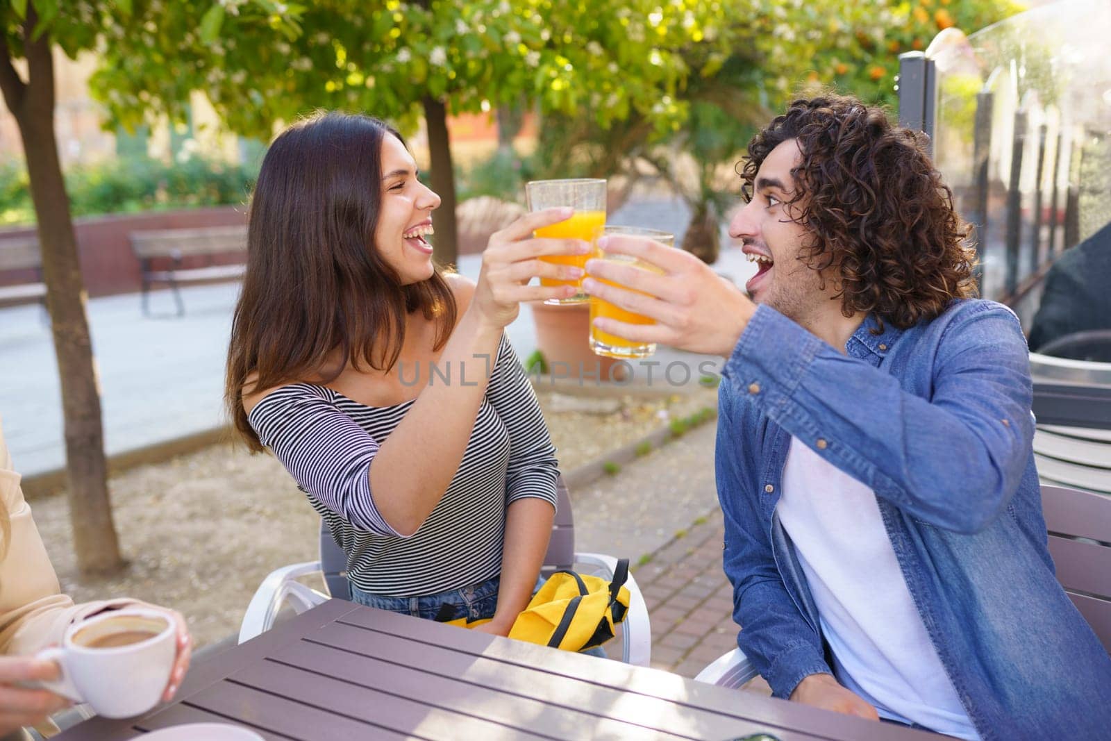 Delighted male and female best friends, clinking glasses with orange juice and celebrating meeting in summer cafe while sitting at table and looking at each other