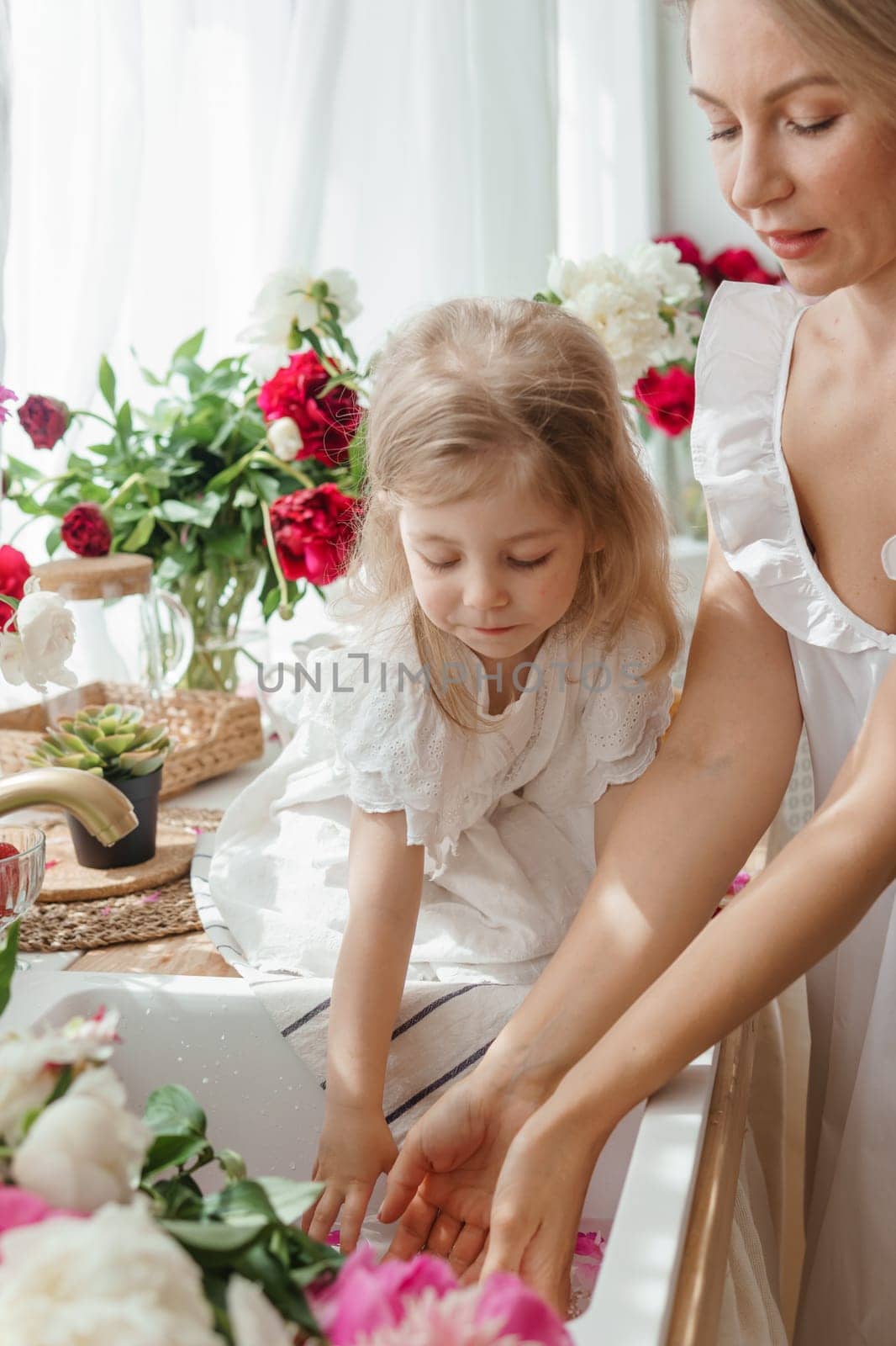 A little blonde girl with her mom on a kitchen countertop decorated with peonies. The concept of the relationship between mother and daughter. Spring atmosphere.